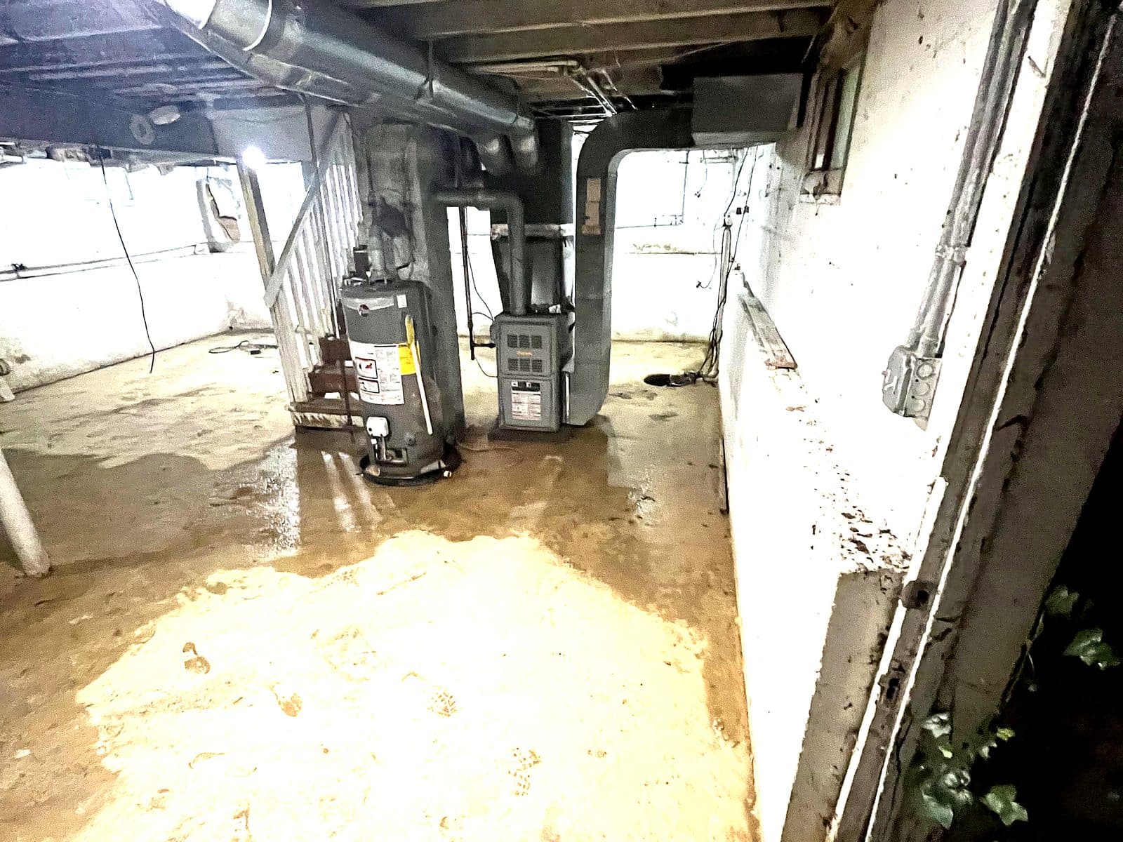Basement of a Maryland home with an aging water heater, old furnace, exposed ductwork and a damp concrete floor